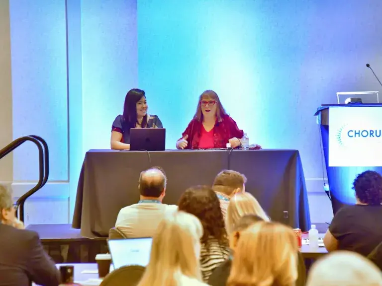 Photo depicts Maggie Vo and Beth Kanter seated side by side behind a table. They are on stage presenting to the audience in front of them